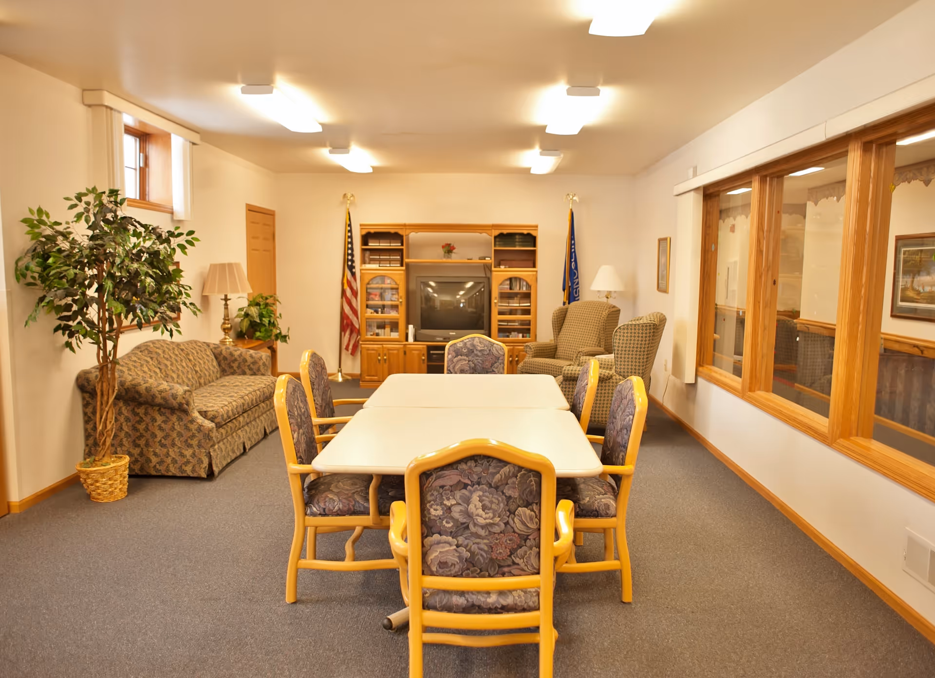 A well-lit common room with a rectangular table surrounded by six floral-patterned chairs in the center. To the left, there is a patterned sofa next to a potted plant and a lamp. At the back of the room, there is a wooden entertainment center with a television, flanked by two flags. To the right, there are two cushioned armchairs and a lamp, with large windows along the right wall showing another room.