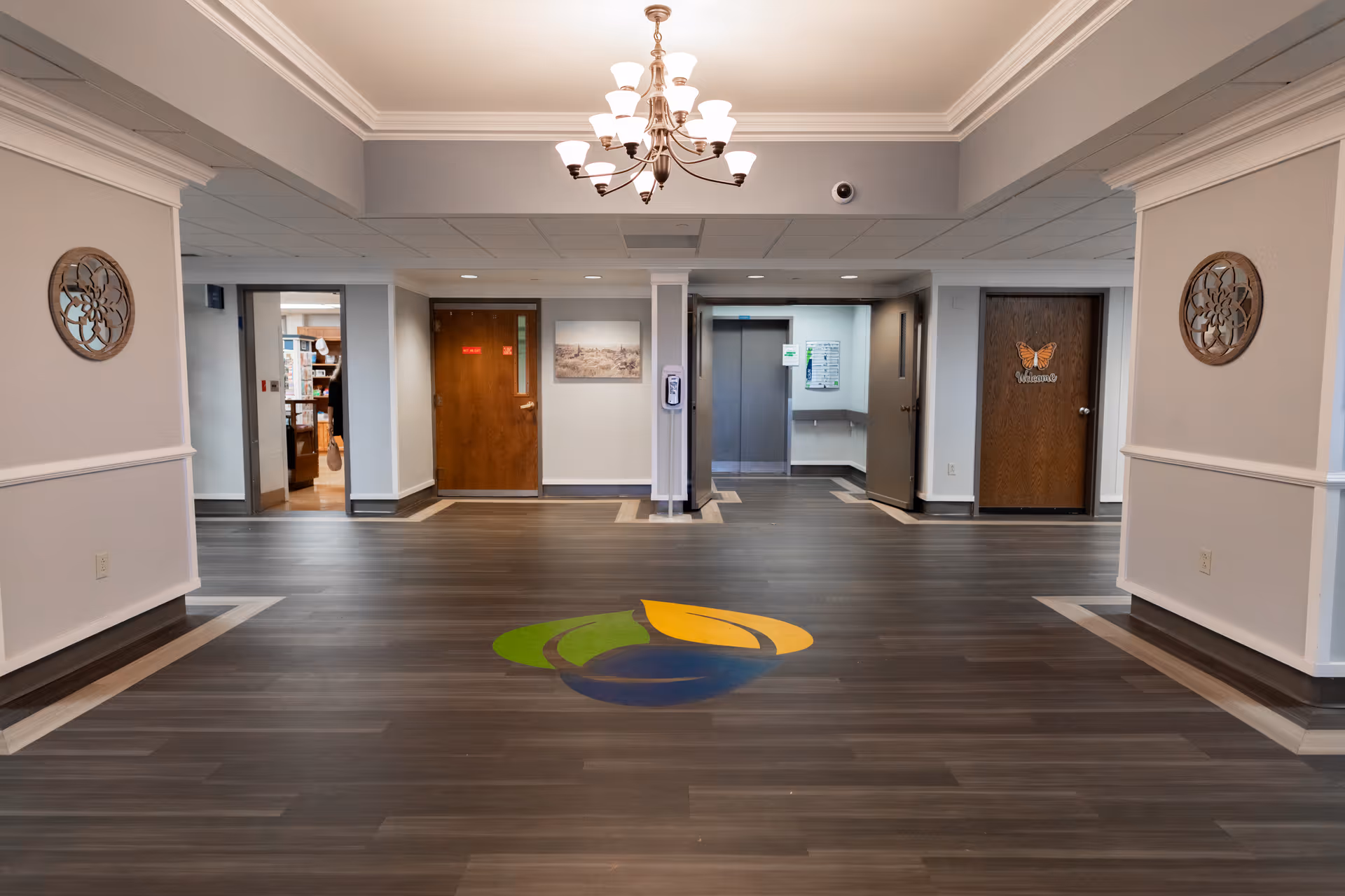 Interior view of a hallway in Masonicare Health Center with wood flooring featuring a colorful leaf logo in the center. The hallway has a chandelier hanging from the ceiling, two wooden doors on the far wall, one with a butterfly decoration and the word 'Welcome'. There is an elevator door in the middle and decorative wall accents on both sides.