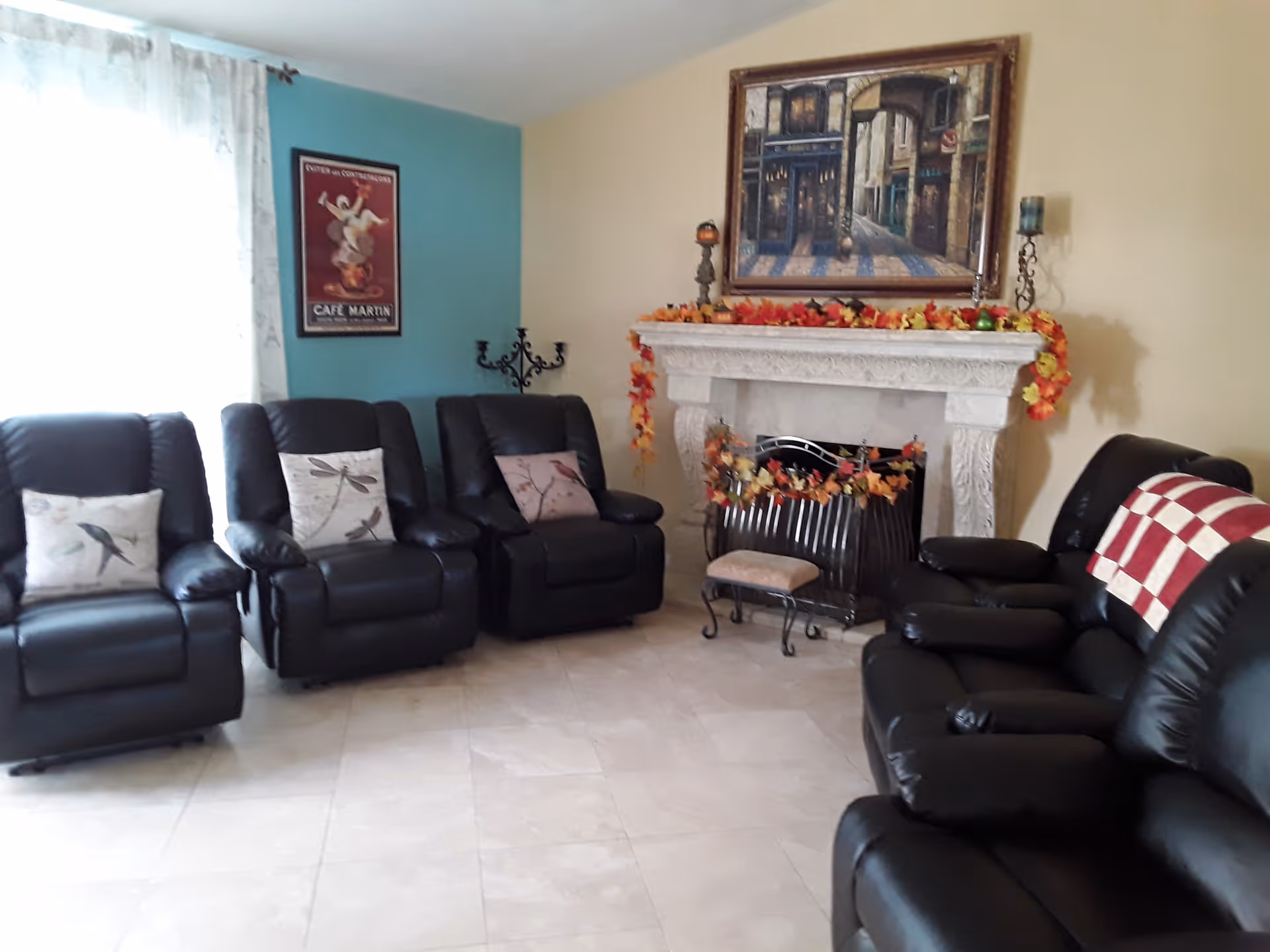 A cozy living room with four black leather recliners arranged along two walls. The room features a decorative white fireplace adorned with autumn-themed garlands and a framed painting above it. There is a window with sheer curtains on the left side and a framed poster on a teal accent wall.