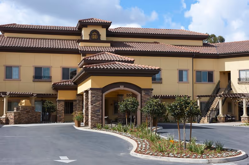 Front entrance and driveway of a two-story Mediterranean-style senior living building with tiled roofs and a landscaped circular island.
