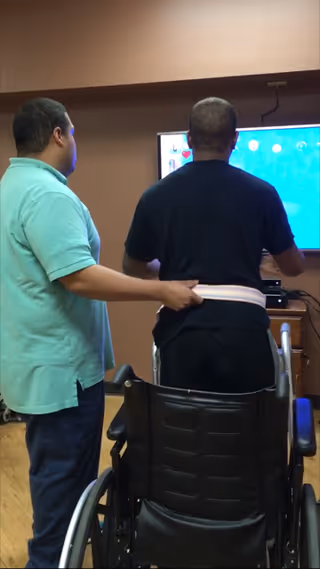 A caregiver assists a man standing in front of a television screen. The man is supported with a gait belt around his waist and a wheelchair is positioned behind him. The setting appears to be an indoor room with wooden flooring and brown walls.