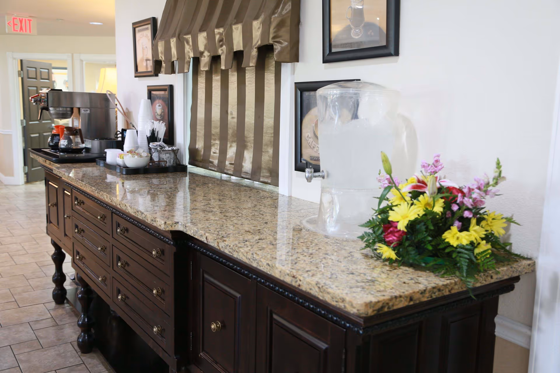 Granite-topped sideboard with a coffee station, water dispenser, and a floral arrangement in a facility interior hallway.