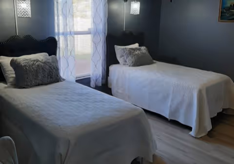 A bedroom with two twin beds covered in white bedspreads, each with a gray decorative pillow. The room has dark walls, a window with sheer white curtains letting in natural light, and two wall-mounted light fixtures above the beds. There is a framed picture on the wall to the right.