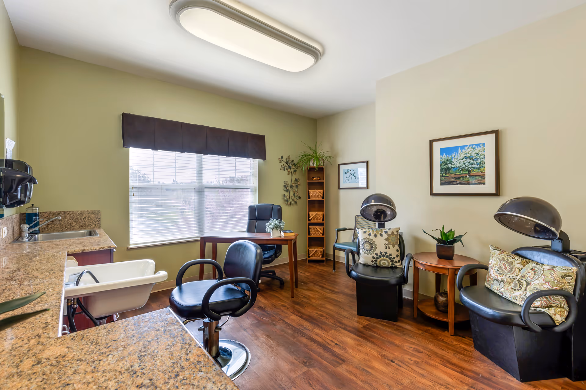 A bright hair salon room with two black hair drying chairs with patterned cushions, a small wooden side table with a plant, a wooden desk with a black office chair, a tall shelving unit with baskets and a plant, a large window with blinds and a dark valance, and a countertop with a sink and a salon chair in front of it. The room has light green and beige walls and wood flooring.