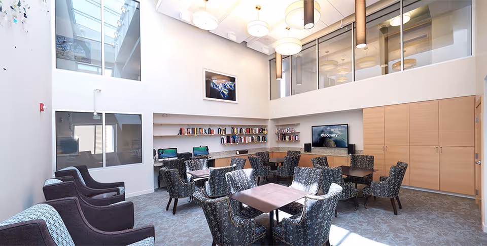 A bright, modern common area with multiple tables and patterned chairs arranged for group seating. The room features high ceilings with large windows and hanging light fixtures. There are bookshelves with books and two computer stations along one wall, and a flat-screen TV mounted on another wall. Light wood cabinets and a door are visible on the right side.