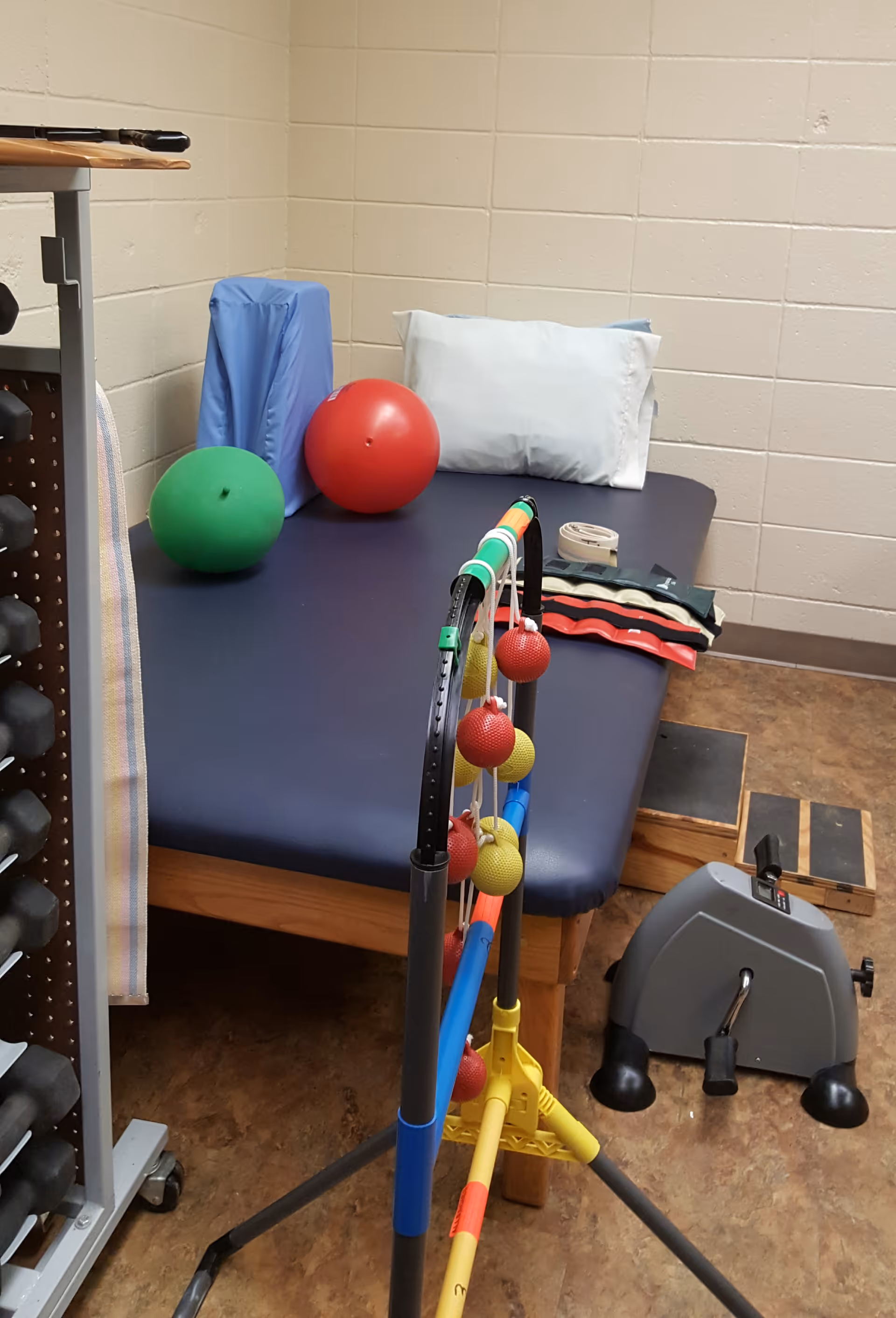 A physical therapy room with a padded treatment table holding a pillow, two exercise balls (one red and one green), and some resistance bands. In front of the table is a ladder toss game with red and yellow bolas hanging. To the right is a small pedal exerciser and a wooden step platform. A rack of dumbbells is partially visible on the left side.