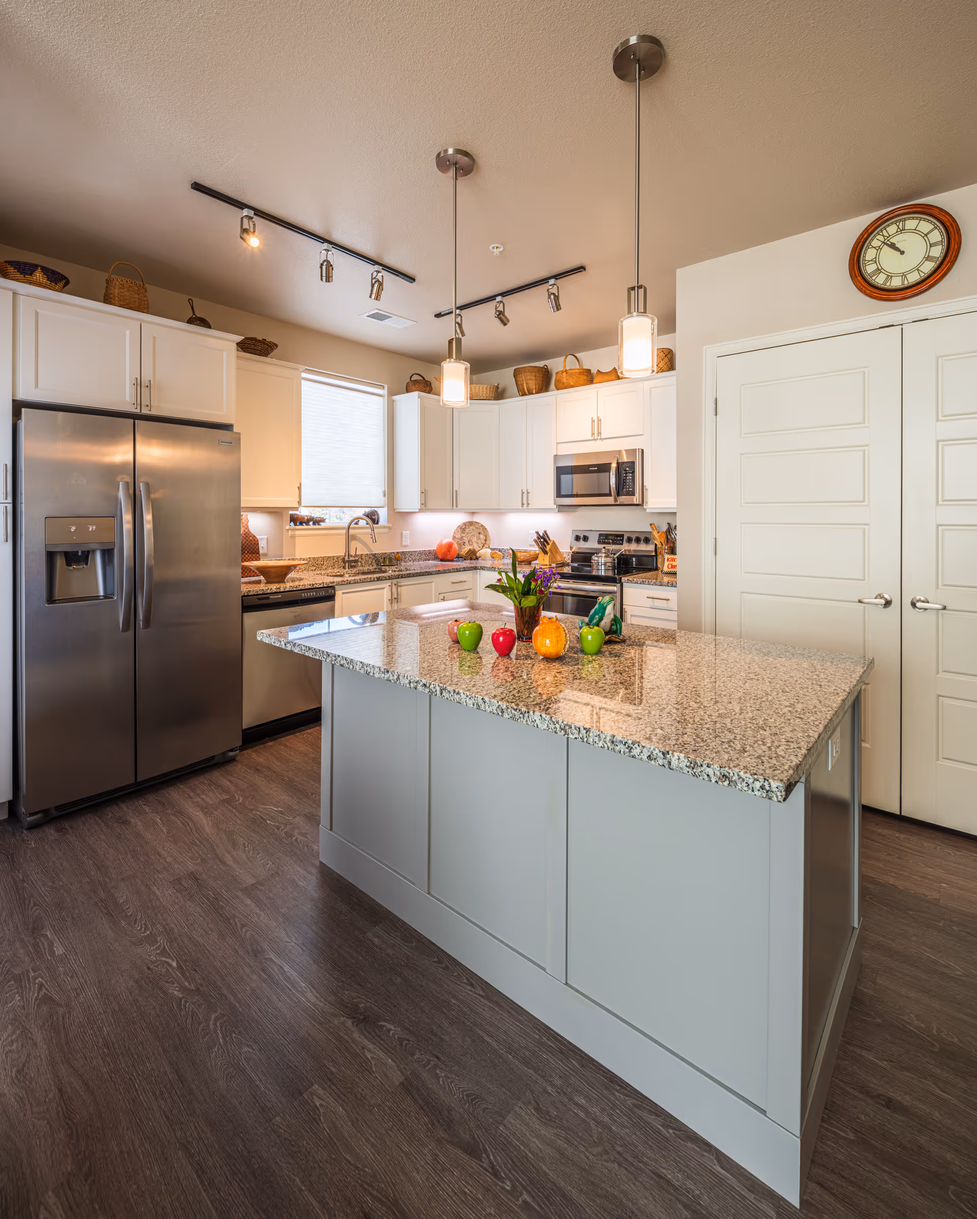 Modern kitchen with a large granite island countertop in the center, decorated with small colorful apple-shaped ornaments and a small plant. The kitchen features white cabinets, stainless steel refrigerator, dishwasher, microwave, and stove. There are pendant lights hanging above the island and a wall clock above double doors on the right side.