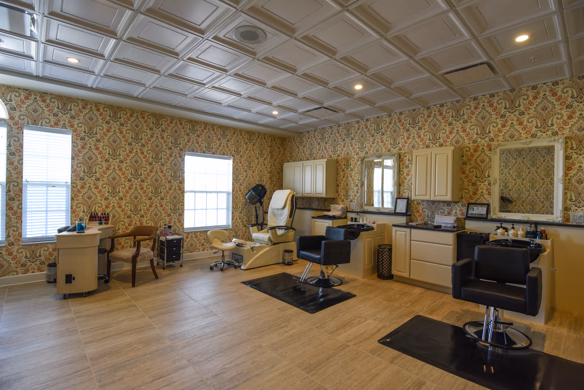 A spacious beauty salon area with patterned wallpaper, two large windows with blinds, and a coffered ceiling. The room contains salon chairs, a pedicure chair, manicure station, mirrors, cabinets, and various salon equipment.