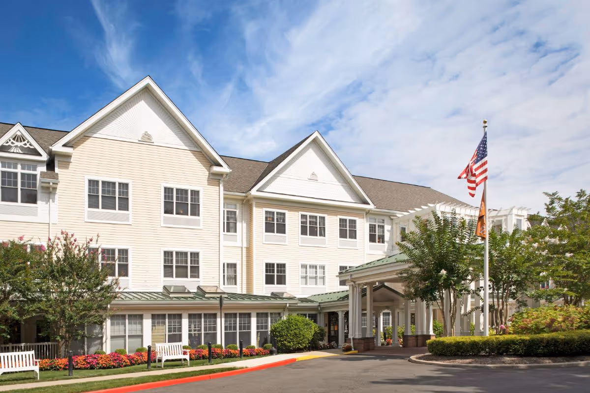 Front exterior of a three-story senior living facility with a covered entrance, American flag, and landscaped grounds.