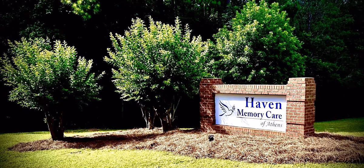 A brick sign for Haven Memory Care of Athens surrounded by green bushes and grass with a wooded background.