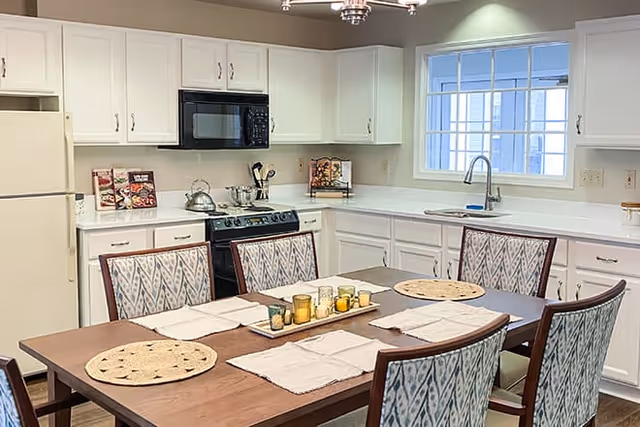 A bright kitchen with white cabinets, a white refrigerator, a black microwave, and a black stove. A wooden dining table with six patterned chairs is set with placemats and a centerpiece of candles. A window above the sink lets in natural light.