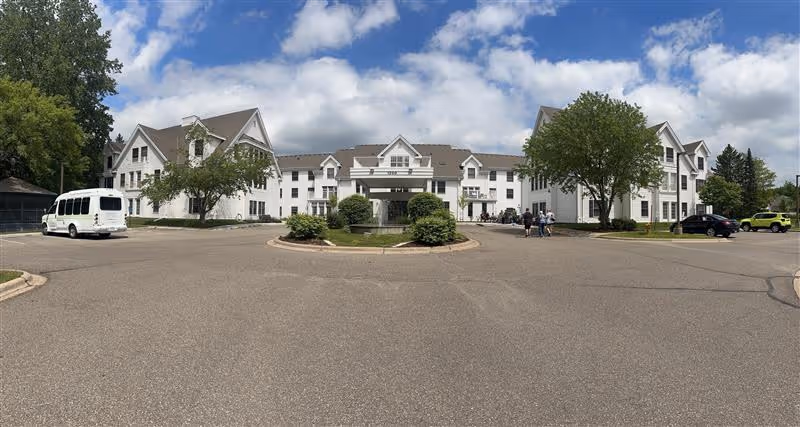 Front exterior of a large white multi-story senior living building with a circular driveway, parked vehicles, and a few people under a partly cloudy sky.