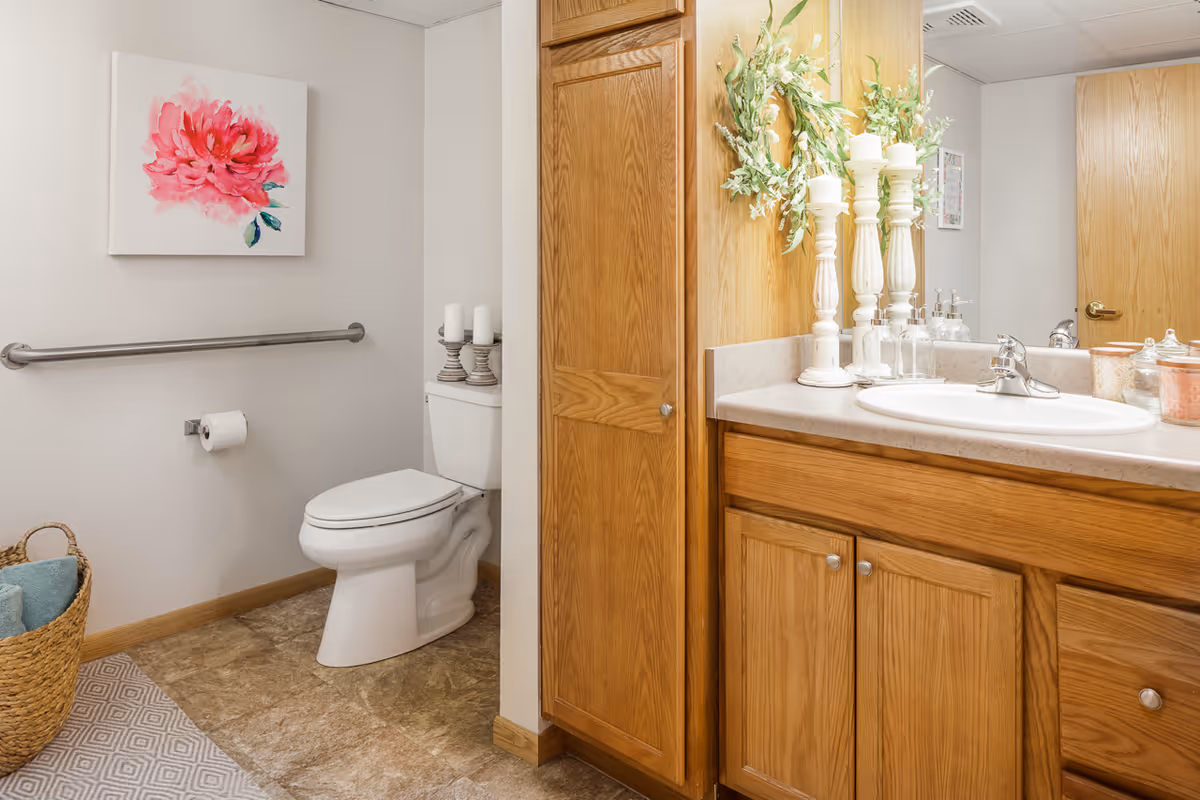 A clean bathroom with a white toilet, a wooden vanity with a sink, and a large mirror. The walls are light-colored with a pink flower painting above a metal grab bar next to the toilet. There is a woven basket with towels on the floor and decorative candles on the toilet tank and vanity.