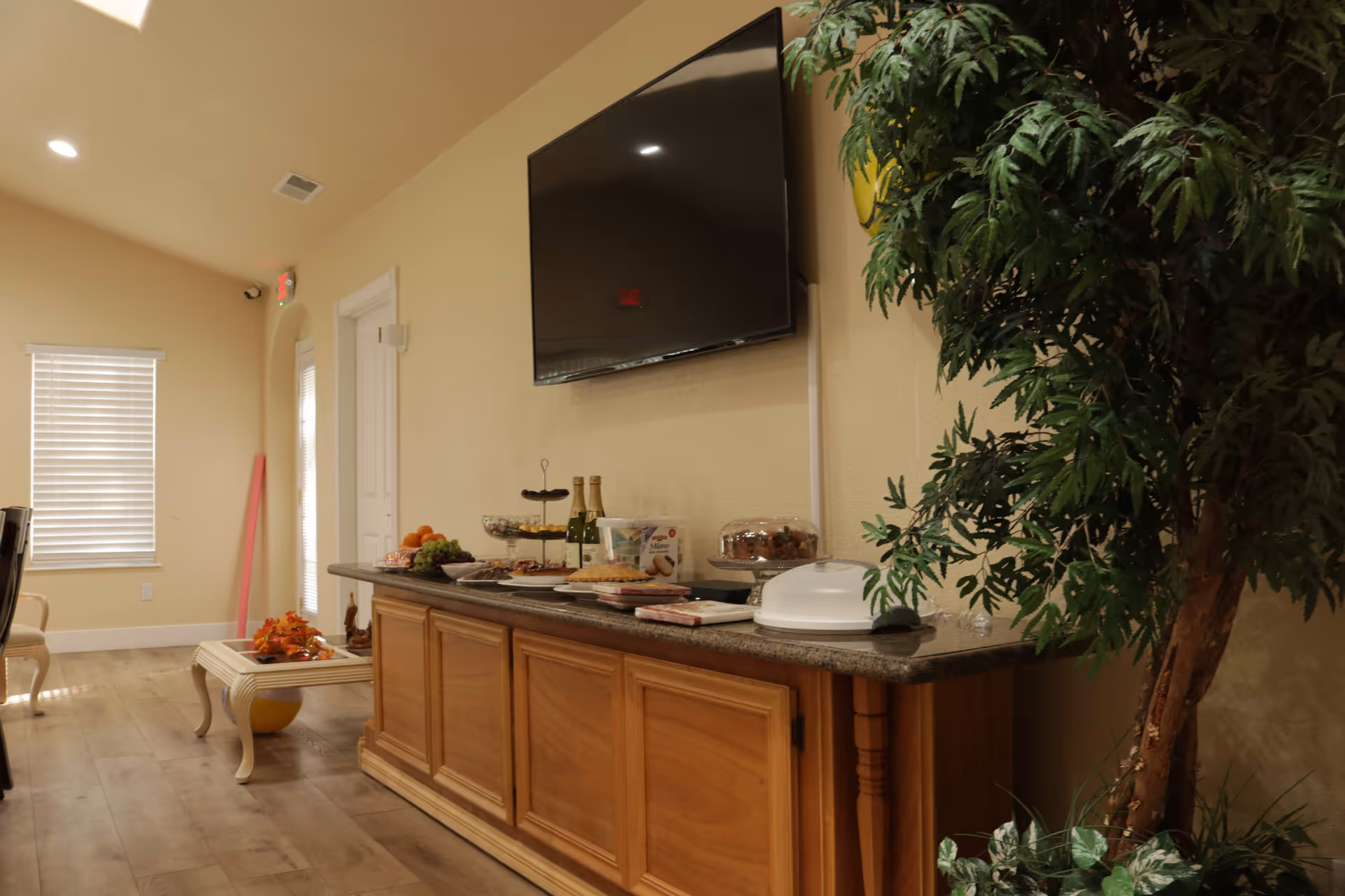 Interior view of a senior care facility room with a wooden cabinet topped with various food items and drinks. A large flat-screen TV is mounted on the beige wall above the cabinet. To the right, there is a large green potted plant. The room has wooden flooring, a window with blinds, and a small table with decorative items near the window.