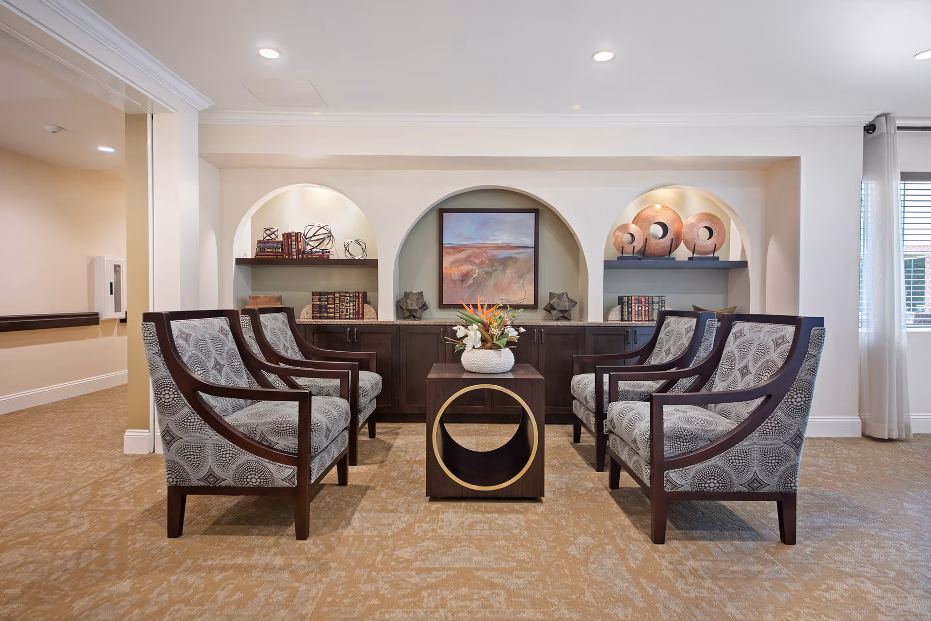A cozy seating area with six patterned armchairs arranged around a small wooden table with a decorative vase of flowers. Behind the chairs is a built-in cabinet with three arched niches displaying books, decorative objects, and a framed painting. The room has beige walls, carpeted floor, and a window with sheer curtains on the right side.