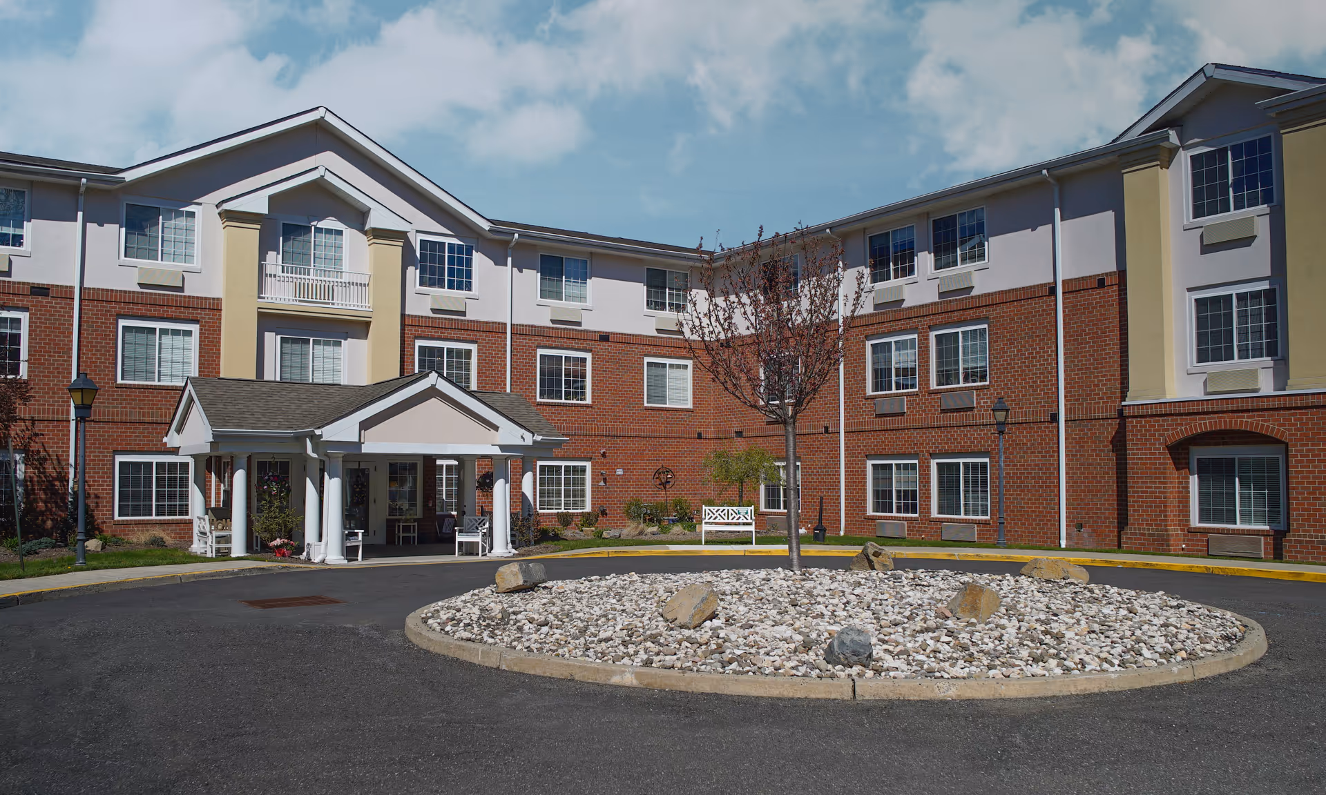 Exterior view of a three-story senior living facility building with a brick and beige facade, multiple windows, a covered entrance with white columns, a circular driveway with a rock and tree centerpiece, and a white bench near the entrance.