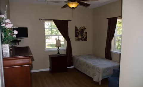 A small bedroom with a single bed covered with a patterned bedspread, two windows with dark curtains, a wooden nightstand with a lamp, a wooden dresser with a vase of flowers, a wall-mounted TV, and a ceiling fan with a light fixture.