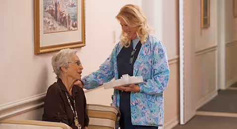 A caregiver in a blue patterned jacket is smiling and interacting with an elderly woman seated on a beige chair in a hallway. The caregiver is holding a tray with cups, and there is a framed painting on the wall behind them.