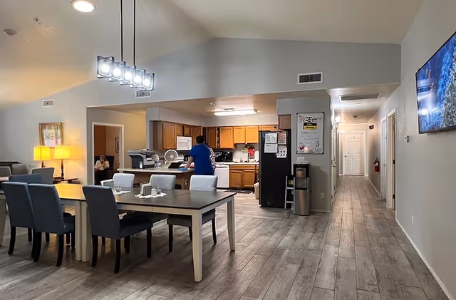 Interior view of a senior living facility showing a dining area with a table and chairs, a kitchen with wooden cabinets and appliances, a person standing near the kitchen counter, and a hallway leading to other rooms. The space is well-lit with modern lighting fixtures and has a light-colored wood floor.