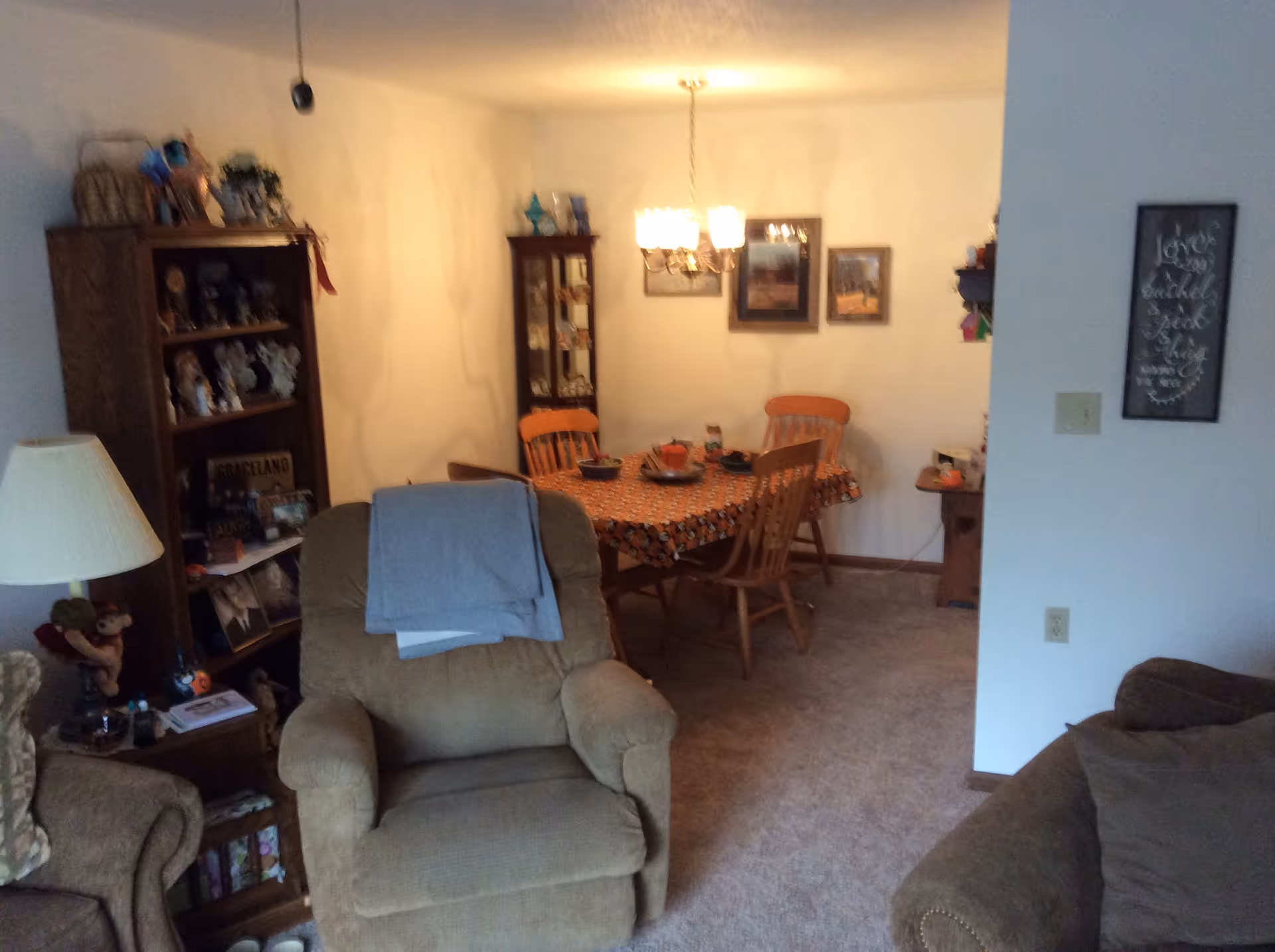 A cozy interior space featuring a beige recliner chair with a folded blanket on the back, a wooden dining table covered with a patterned tablecloth and surrounded by wooden chairs, a wooden cabinet filled with decorative items, a floor lamp, and framed pictures on the walls. The room has carpeted flooring and warm lighting from a chandelier above the dining table.