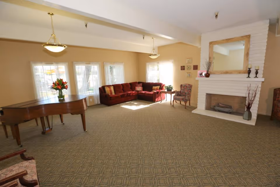 A spacious living room with beige walls and carpeted floor featuring a red sectional sofa with patterned pillows, a floral armchair, a wooden grand piano with a vase of flowers on top, and a white brick fireplace with a large wooden-framed mirror above it. The room is well-lit by natural light coming through multiple windows with sheer curtains.