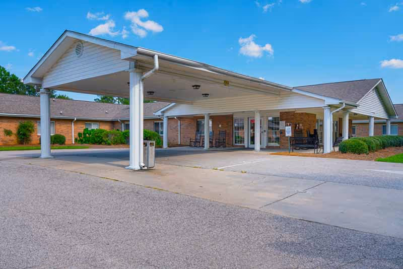 Exterior view of Wilson Healthcare and Rehabilitation Center showing a covered entrance with white pillars, a driveway, and a brick building under a blue sky with some clouds.