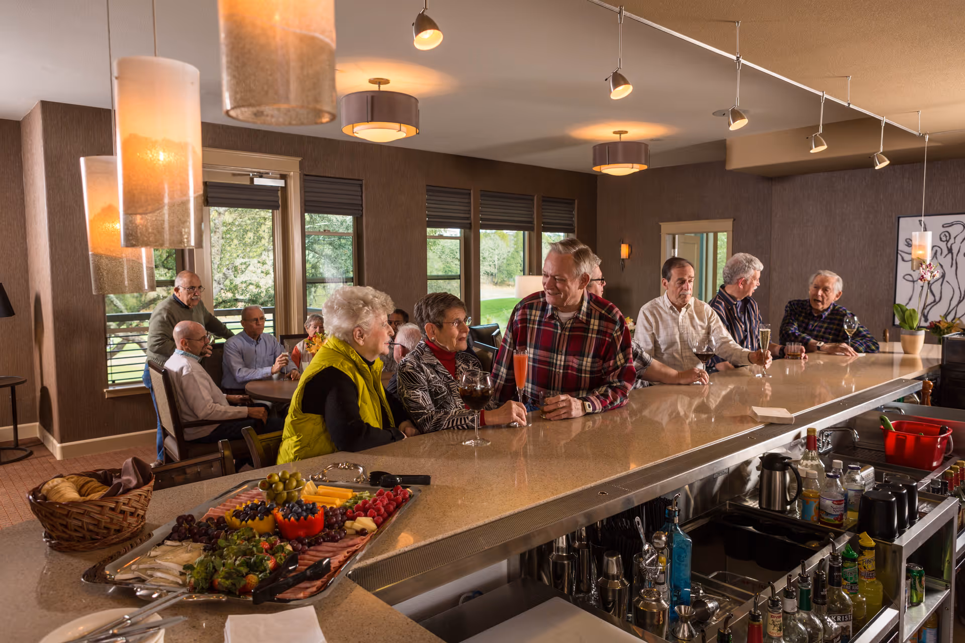 A group of elderly people socializing at a large bar counter inside a cozy room with large windows showing greenery outside. The bar counter has a platter of assorted fruits and snacks, and various bottles and glasses are visible behind the counter. Warm lighting fixtures hang from the ceiling, creating a welcoming atmosphere.