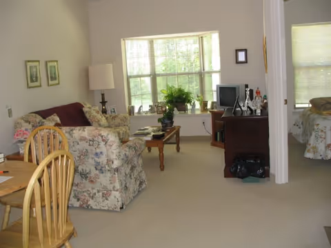 Sunlit open living room with a floral sofa, coffee table, TV stand and dining table, with a bedroom visible through an open doorway.