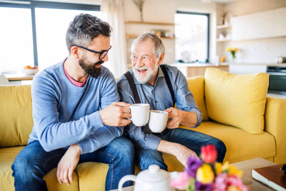An elderly man and a younger man sitting on a yellow couch in a bright living room, smiling and clinking white coffee mugs together. There is a coffee pot and a bouquet of colorful flowers on the table in front of them.