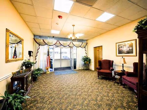 Nursing facility lobby with patterned carpet, seating, plants, draped windows, flag, and entrance doors.