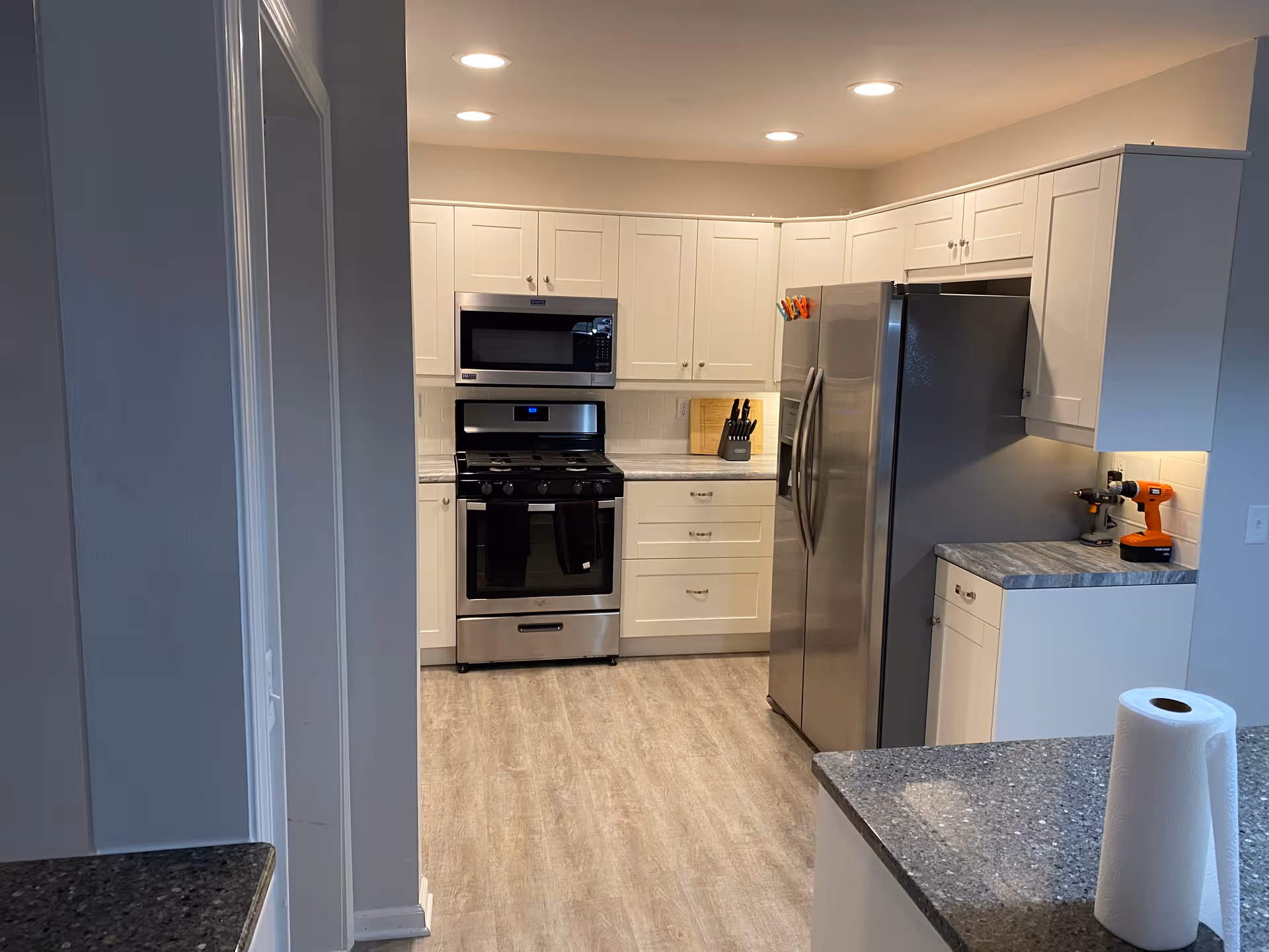Modern kitchen with white cabinets, stainless steel refrigerator, stove, and microwave. The kitchen has light wood flooring and gray countertops. A roll of paper towels is on the counter in the foreground, and a cordless drill is on the counter near the refrigerator.