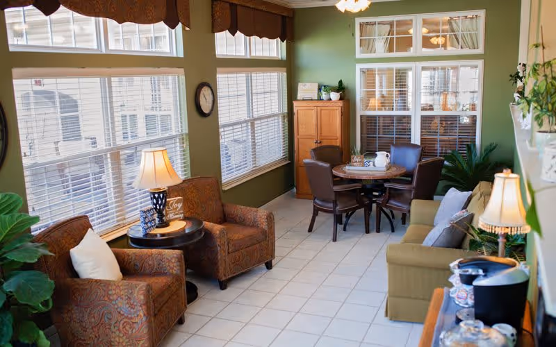 A cozy senior living community common area with green walls and large windows letting in natural light. The room features two patterned armchairs with a small round table and lamp between them, a round table with four brown chairs, a wooden cabinet, and a green sofa with pillows. There are plants and decorative items around the room, creating a warm and inviting atmosphere.