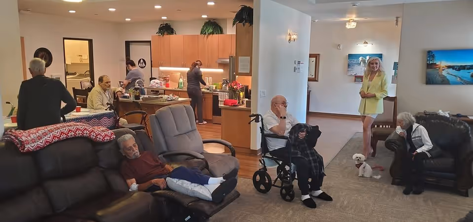 A common area in a senior living facility with several elderly residents and a caregiver. One elderly man is sitting in a wheelchair, another is resting on a recliner, and two others are seated on couches. A caregiver in a yellow outfit stands near a small white dog. In the background, two staff members are working in the kitchen area.