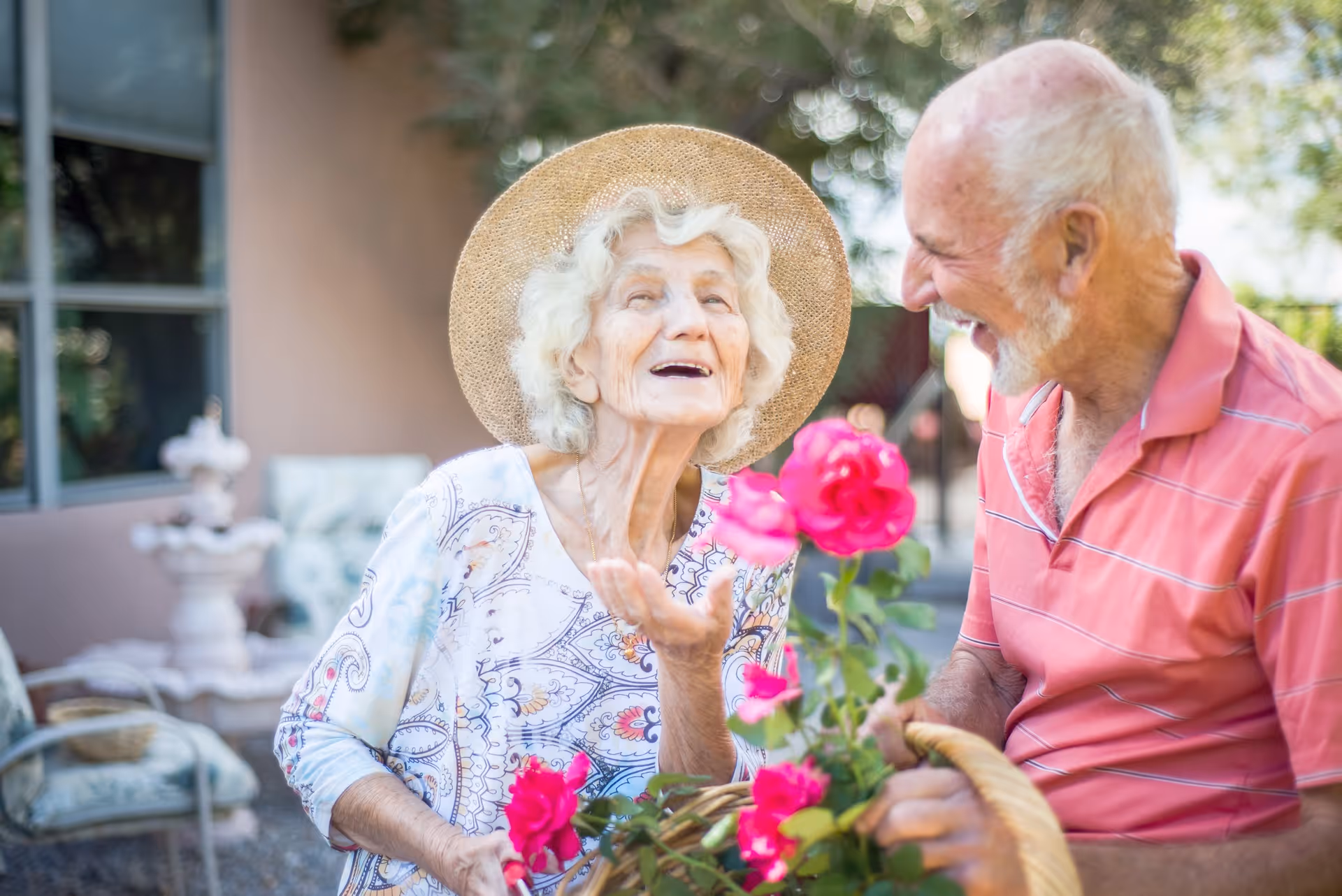 An elderly woman wearing a straw hat and a patterned blouse is smiling and gesturing while holding a basket with pink flowers. An elderly man in a pink striped polo shirt is smiling at her. They are outdoors with greenery and a building in the background.