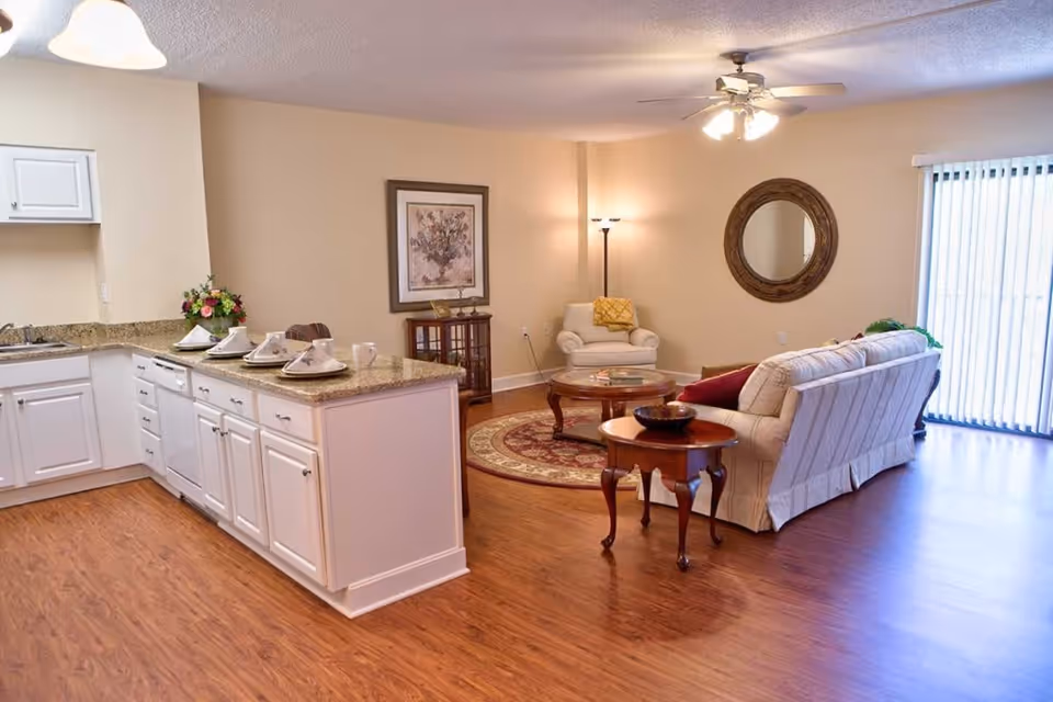 A bright and spacious living area in a senior living community featuring a kitchen with white cabinets and granite countertops on the left, set with plates and cups. The living room area has a beige sofa, an armchair, a round wooden coffee table on a patterned rug, a floor lamp, a round decorative mirror on the wall, and vertical blinds covering a sliding glass door.