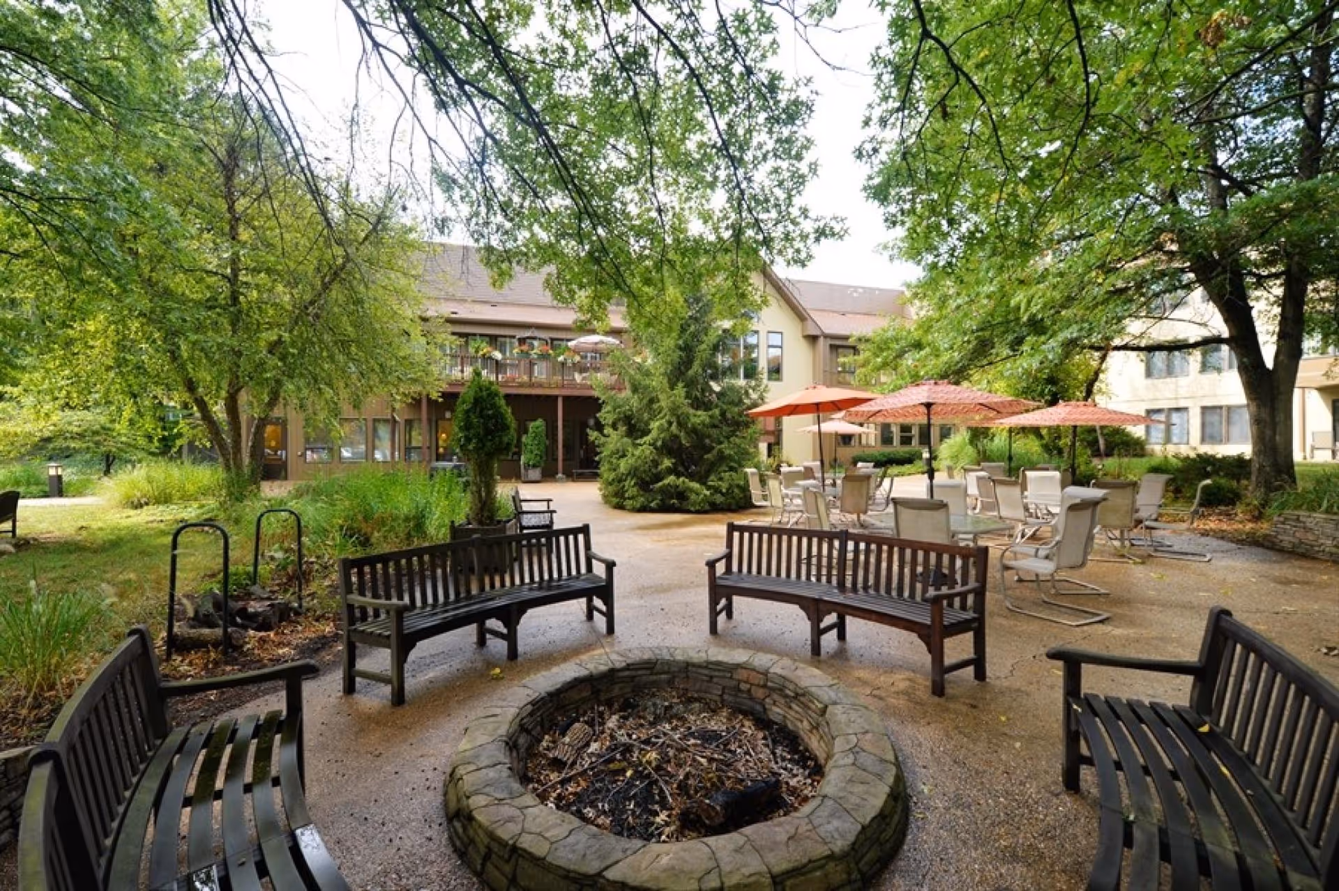 Outdoor courtyard area with a circular stone fire pit surrounded by wooden benches. There are several trees providing shade and a building with balconies and windows in the background. Patio tables with umbrellas and chairs are also visible on the right side.
