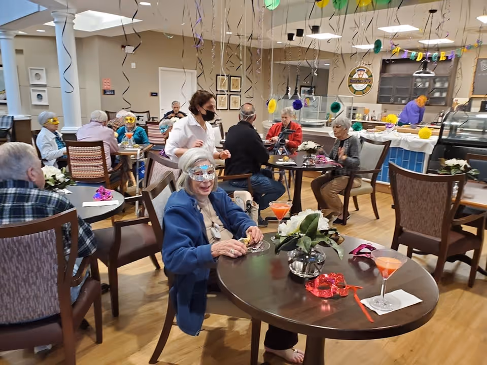 A group of elderly people wearing festive masks and sitting at tables in a decorated dining area. A staff member wearing a mask is attending to them. The room has hanging streamers and colorful decorations, with a counter and kitchen area in the background.
