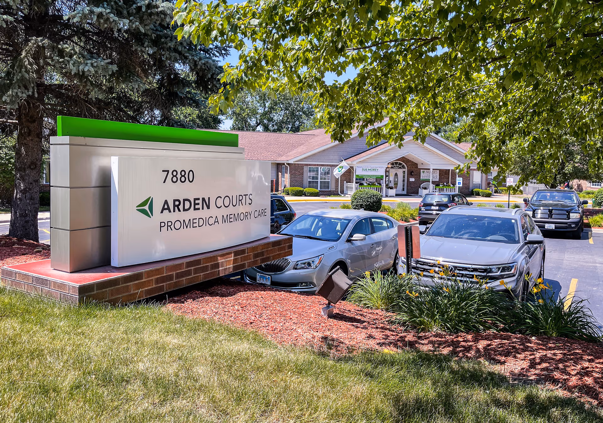 Exterior view of Arden Courts - ProMedica Memory Care Community in Palos Heights, showing a large sign with the address 7880 and the facility name. Several cars are parked in the lot, and the building entrance is visible in the background surrounded by trees and landscaping.