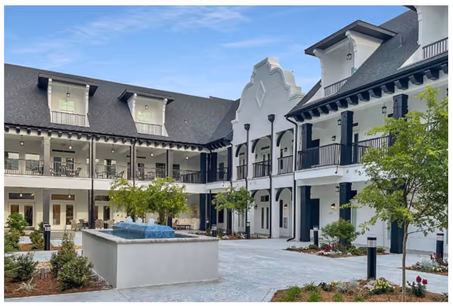 Exterior courtyard of The Blake at St. Johns featuring a two-story white building with black trim, balconies, a central fountain, landscaped greenery, and a clear blue sky.