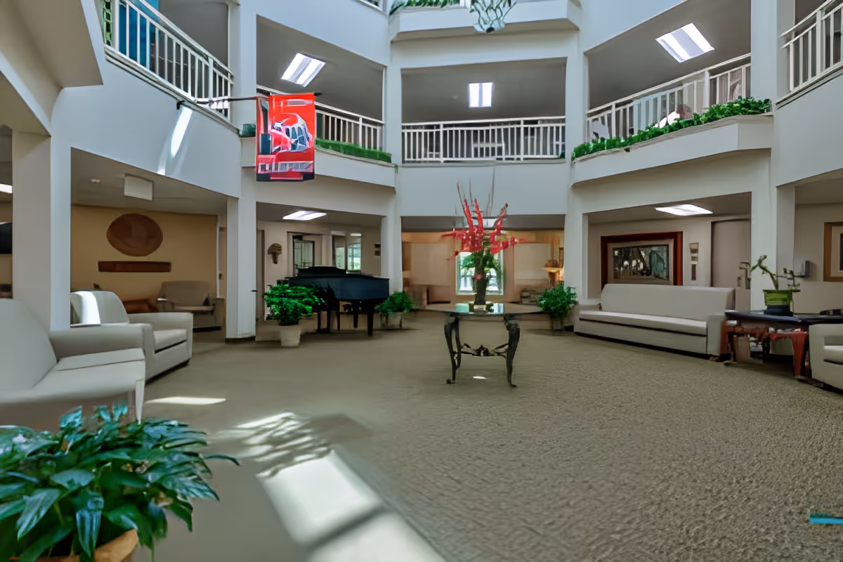Spacious, well-lit common area in a senior living facility with white sofas and armchairs arranged around a central glass table holding a vase with red flowers. There is a black grand piano in the background, potted plants, and a two-story open atrium with railings and skylights above.