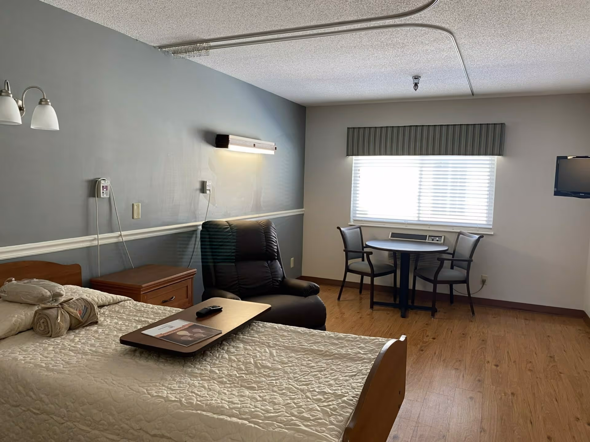 A nursing home bedroom with a single bed covered in a white quilt, a wooden nightstand, a black recliner chair, a small round table with two chairs near a window with blinds, and a wall-mounted TV. The room has wooden flooring and gray and white walls.