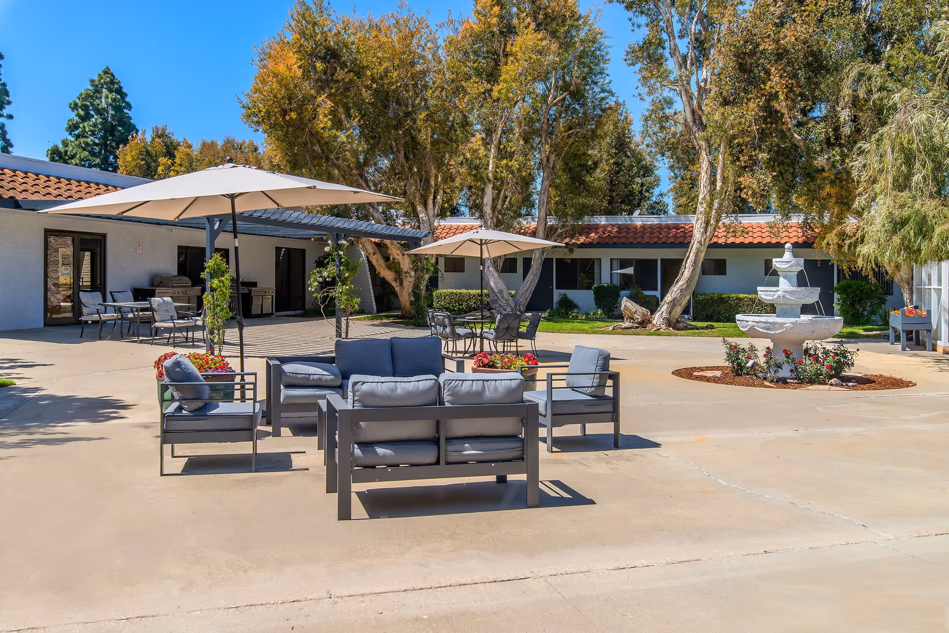 Sunny courtyard at an assisted living facility featuring outdoor lounge seating, umbrellas, a central fountain, and surrounding single-story buildings.