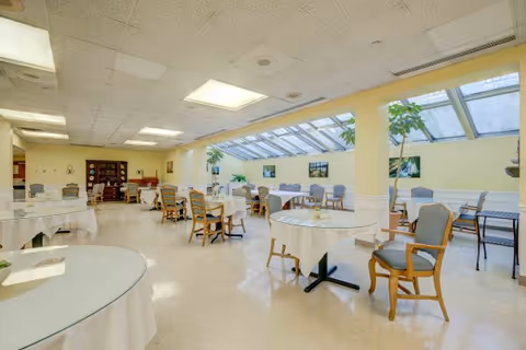 A bright dining room with multiple round tables covered with white tablecloths and surrounded by wooden chairs with blue cushions. The room has large windows and a glass ceiling on one side, allowing natural light to fill the space. There are plants and framed pictures on the walls, creating a welcoming atmosphere.
