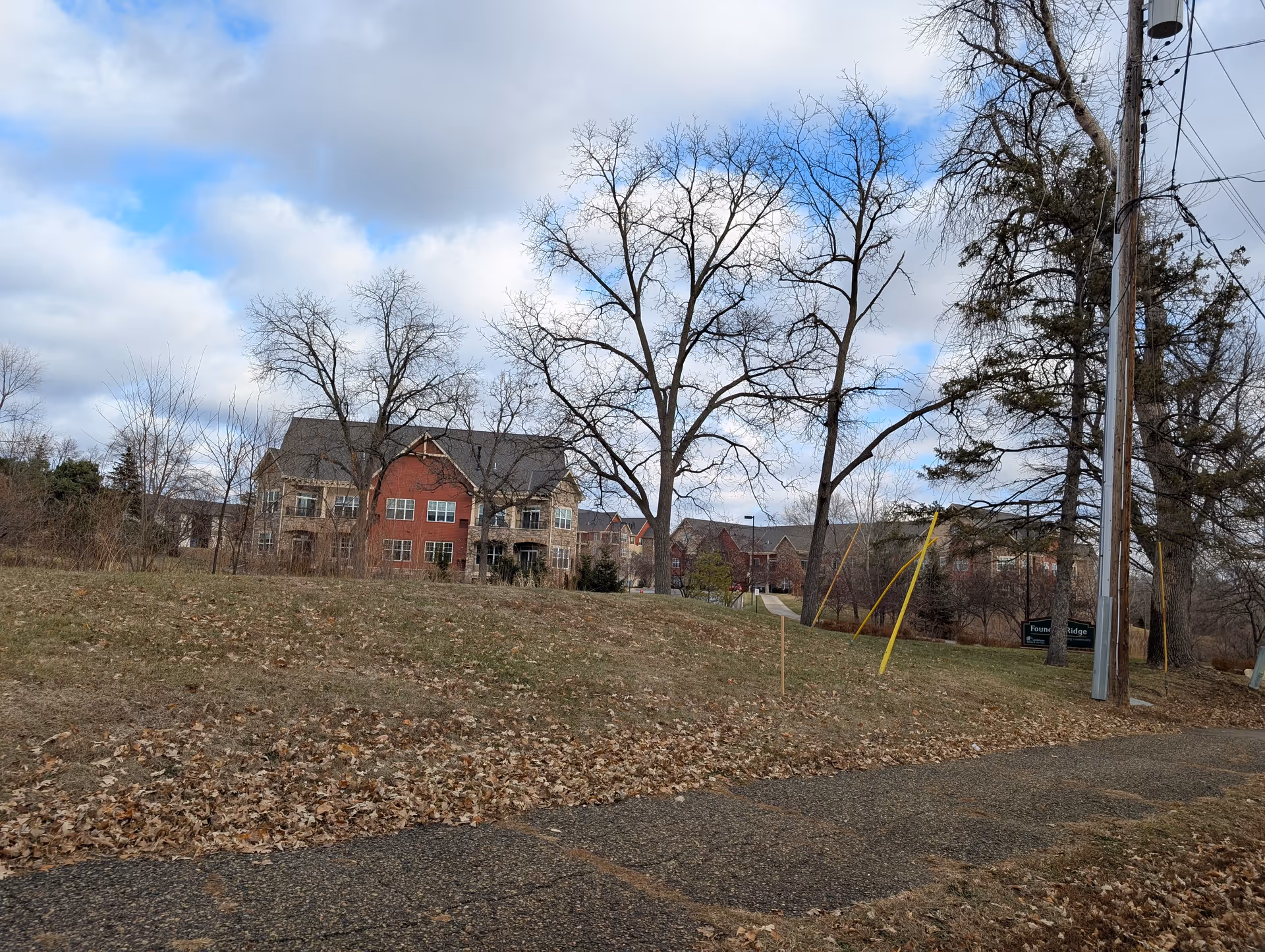 View of a senior living facility named Founders Ridge with multi-story buildings in the background, leafless trees, a grassy area with fallen leaves, a paved pathway, and a partly cloudy sky.