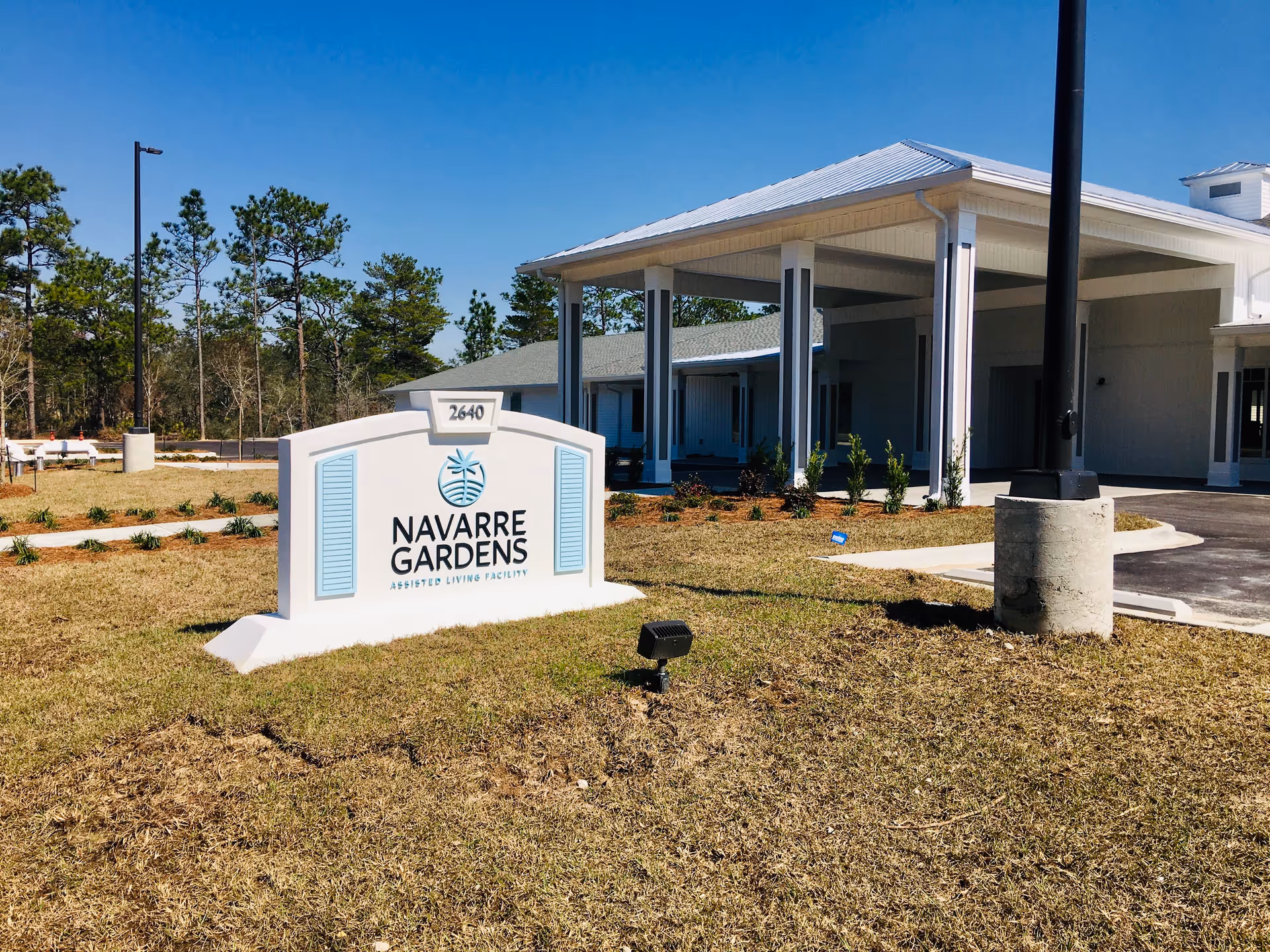Exterior view of Navarre Gardens assisted living facility with a white sign displaying the facility name and address 2640, a covered entrance with pillars, and a clear blue sky.