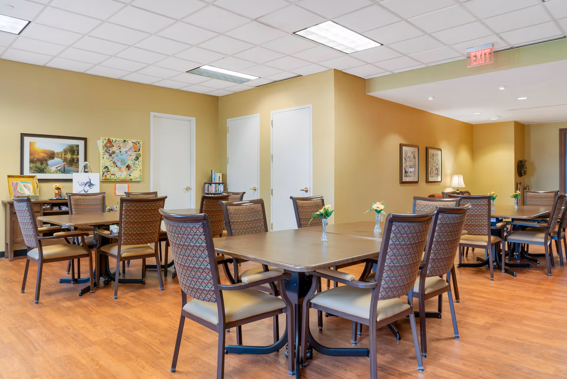 A well-lit dining area with multiple wooden tables and cushioned chairs arranged neatly. Each table has a small vase with yellow flowers. The room has light brown walls, wooden flooring, and framed artwork on the walls. There are two white doors and an exit sign visible on the ceiling.