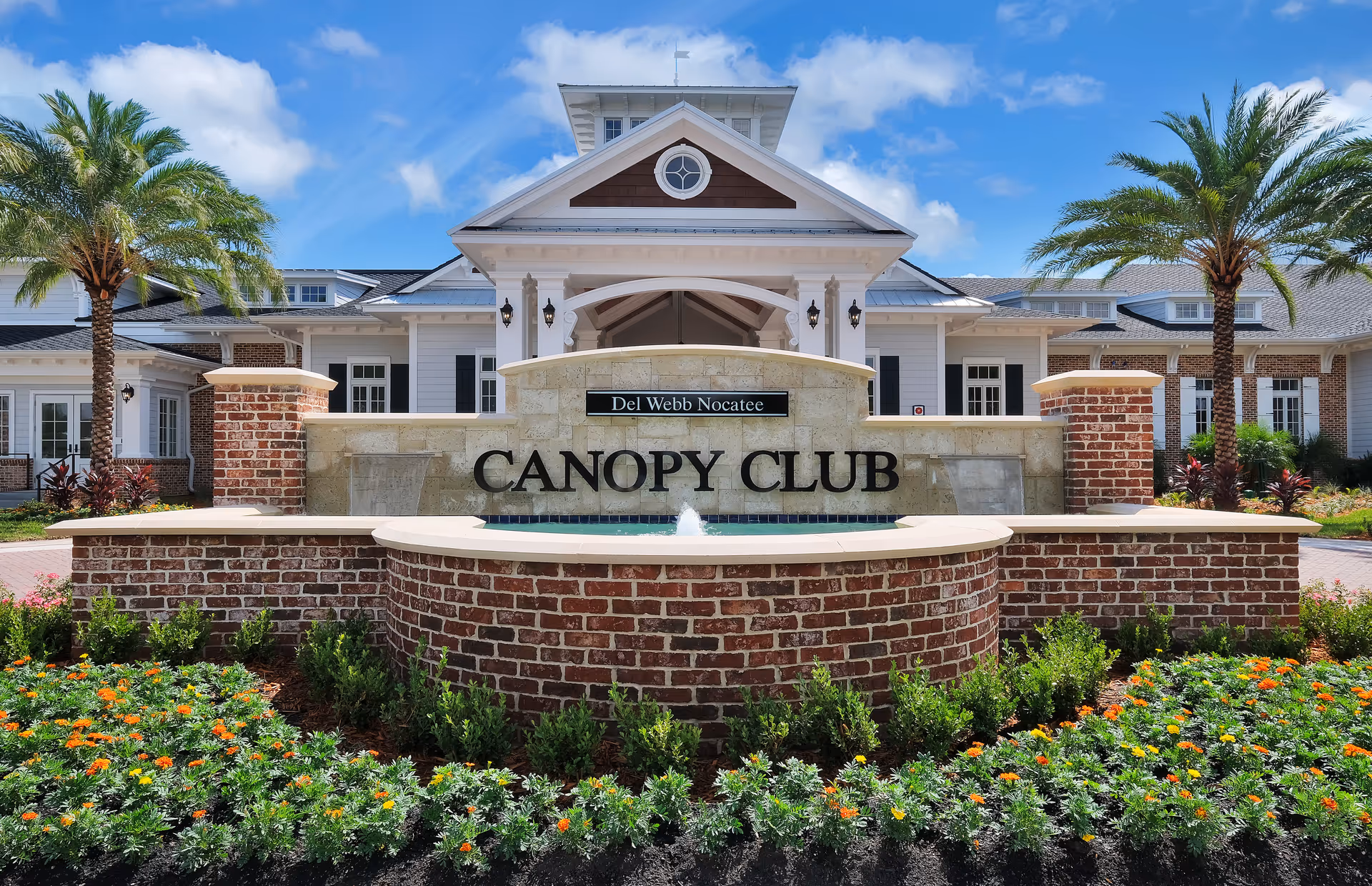 Front entrance of the Canopy Club at Del Webb Nocatee with a brick sign and fountain surrounded by landscaped flowerbeds and palm trees.