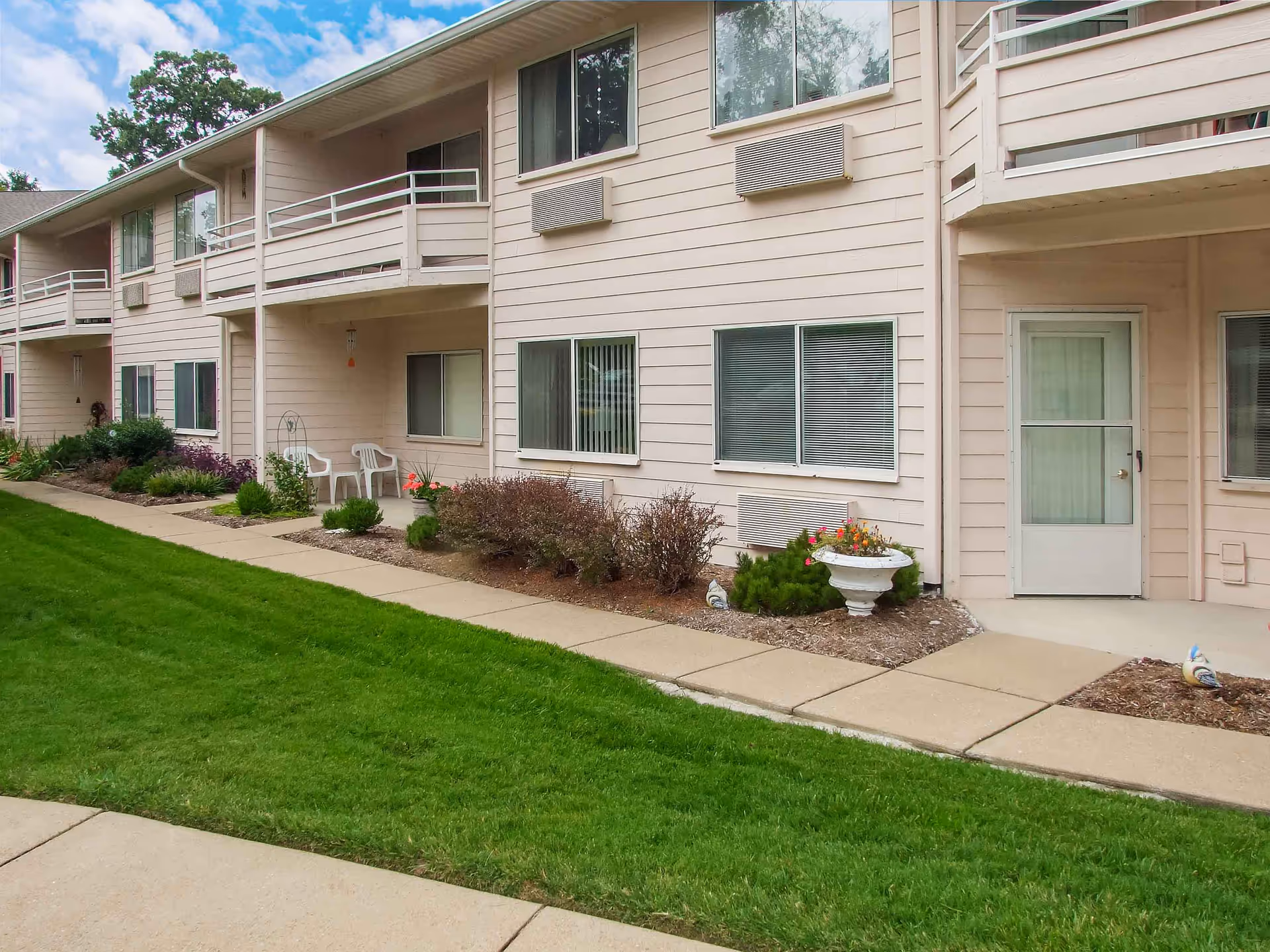 Exterior view of a beige two-story residential building with balconies, windows, and a door. There is a concrete walkway in front of the building, bordered by green grass and small landscaped garden beds with shrubs and flowers. Two white plastic chairs are placed on a small patio area.