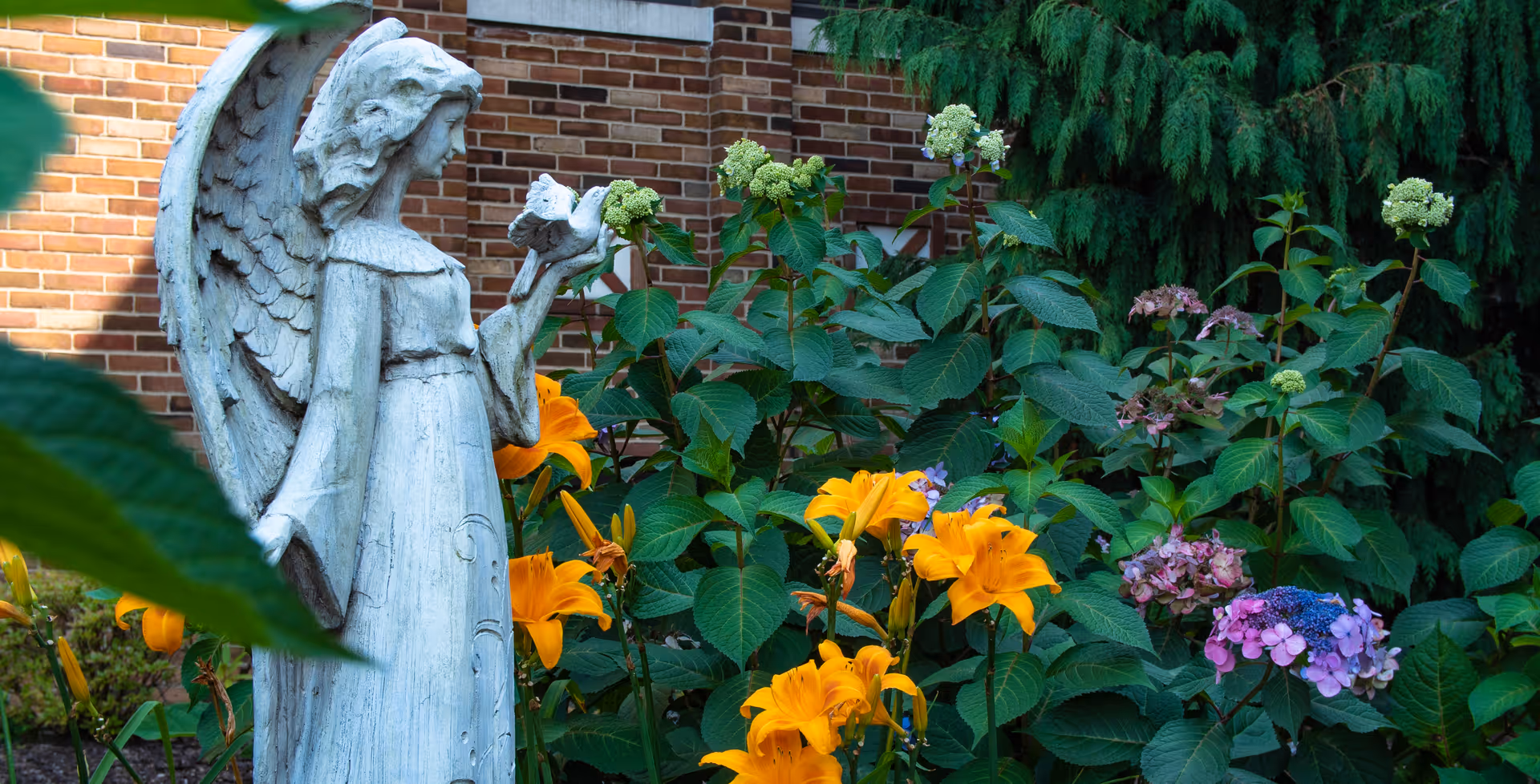 A white stone angel statue holding a bird, surrounded by vibrant orange lilies and purple hydrangeas in a garden area with a brick wall and green foliage in the background.