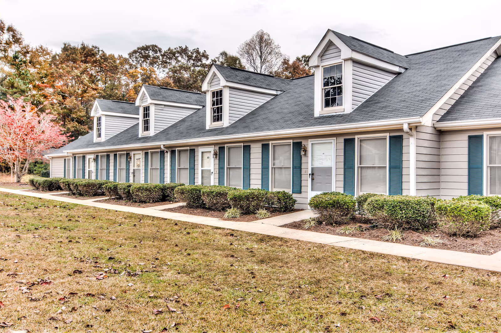 Exterior view of a single-story residential building with multiple windows and doors, blue shutters, and a gray roof with dormer windows. There is a sidewalk in front of the building and neatly trimmed bushes along the walkway. Trees with autumn foliage are visible in the background.